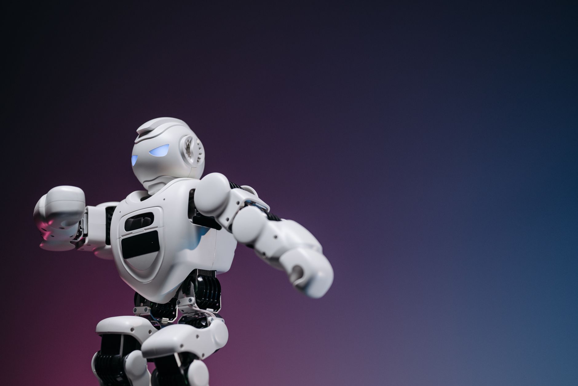 Pequeno robô de brinquedo branco sobre fundo rosa e azul, representando assistentes digitais e robôs de IA.